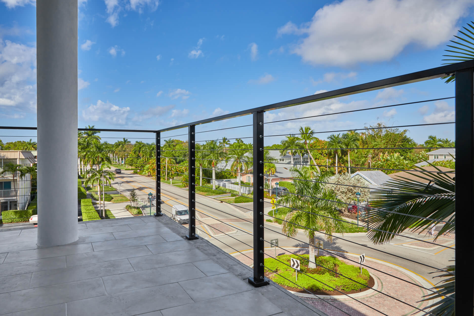 This white deck with metal railings overlooks a street lined with houses and palm trees. A car is entering the round-about.